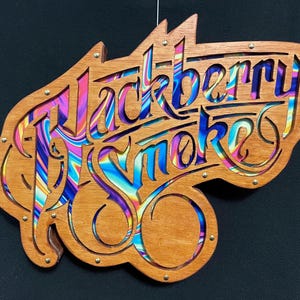 Könnte beinhalten: Ein Holzschild mit den ausgeschnittenen Worten "Blackberry Smoke". Der Hintergrund ist ein farbenfrohes Wirbelmuster.