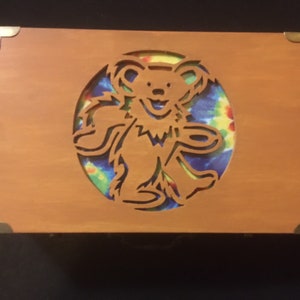 May include: A wooden box with a cutout of a smiling bear in a tie-dye circle. The box has brass corner accents.