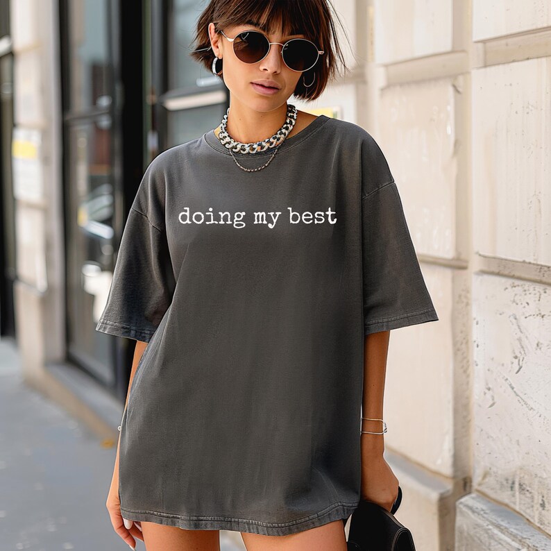 May include: Dark gray t-shirt with the text "doing my best" in white. The shirt has short sleeves and a relaxed fit. The model is wearing sunglasses and a silver chain necklace. The shirt is a casual, everyday wear item.