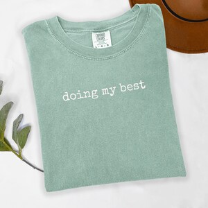 May include: A sage green t-shirt with the phrase "doing my best" in white lettering. The shirt has a crew neck and short sleeves. A brown hat is partially visible in the background.