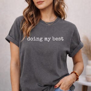 May include: Dark gray t-shirt with rolled-up sleeves. The phrase "doing my best" is printed in white across the chest. The shirt is a solid color and appears to be made of a soft material.