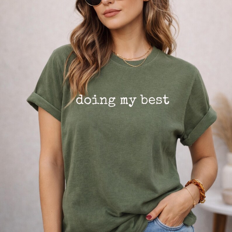 May include: Olive green t-shirt with rolled-up sleeves. The white text "doing my best" is printed across the chest. The model is wearing a gold necklace and a brown bracelet. The shirt is made of a soft, comfortable material.