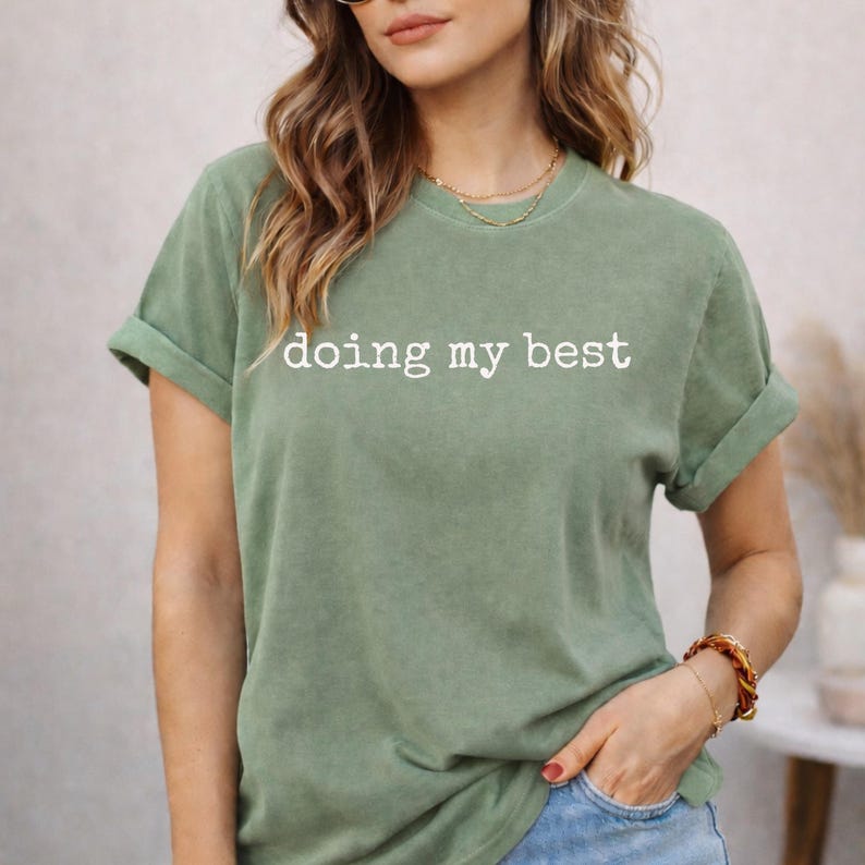 May include: Sage green t-shirt with rolled-up sleeves. The white text "doing my best" is centered on the front. The model is wearing a gold necklace and a bracelet. The shirt is made of a soft, comfortable material.