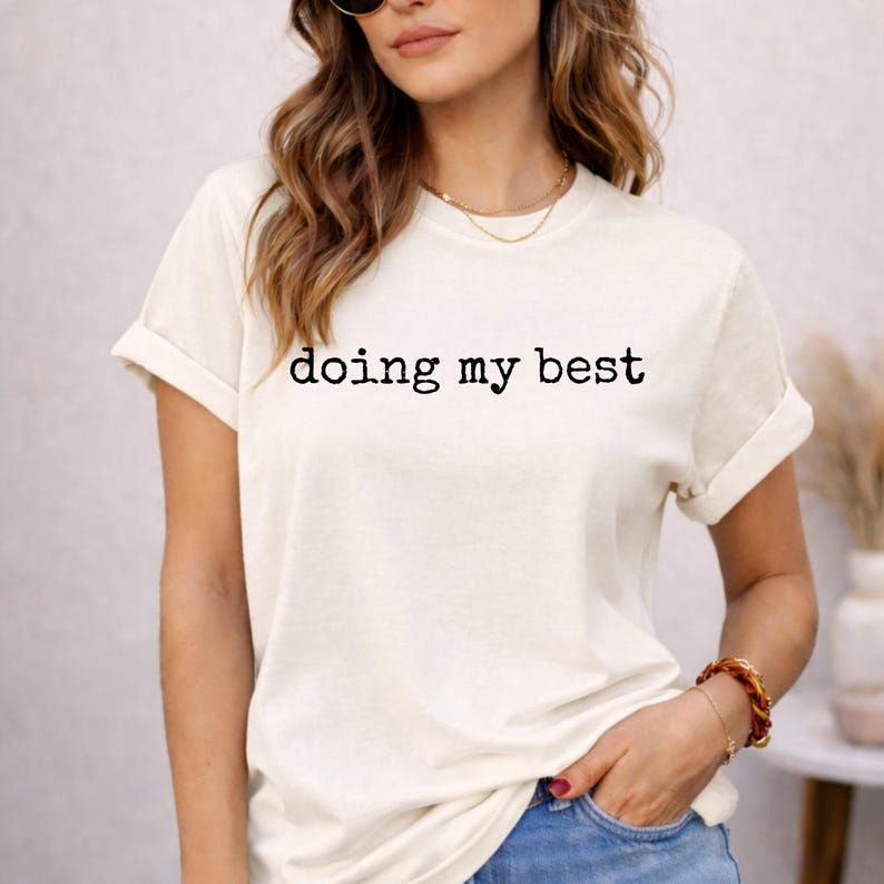 May include: Cream-colored t-shirt with the phrase "doing my best" printed in black. The short-sleeved shirt is rolled up at the sleeves. The model is wearing a gold necklace and a bracelet.
