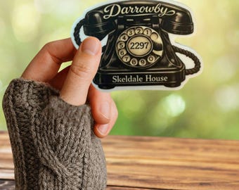 May include: A die-cut sticker featuring a black vintage rotary telephone with the text Darrowby at the top and Skeldale House at the bottom. The centre of the rotary dial displays the number 2297 in a clear, legible font against a cream background.