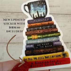 Può includere: Un adesivo raffigurante una pila di titoli di libri di Outlander sormontata da una tazza di notte stellata. L'adesivo include il testo "NEW UPDATED STICKER WITH BOOK 10 INCLUDED!" e titoli di libri come "Voyager" e "Go Tell the Bees That I Am Gone."