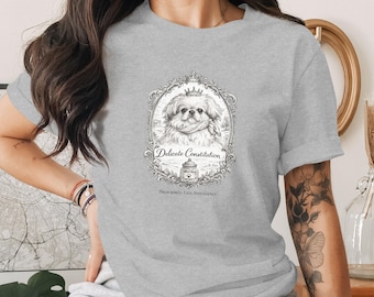 May include: A light grey t-shirt with a black and white illustration of a dog wearing a crown, set in an ornate frame. The text reads "Delicate Constitution" and "Prescribed, Less Indulgence".