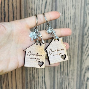 K&ouml;nnte beinhalten: Zwei Schl&uuml;sselanh&auml;nger aus Holz in Hausform mit dem Text "Ci vediamo a casa" (Wir sehen uns zu Hause) und einem Herz-Ausschnitt. Jeder Schl&uuml;sselanh&auml;nger hat einen silbernen Anh&auml;nger, einer mit einer Hantel und der andere mit einem Basketball.