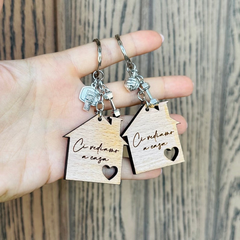 K&ouml;nnte beinhalten: Zwei Schl&uuml;sselanh&auml;nger aus Holz in Hausform mit dem Text "Ci vediamo a casa" (Wir sehen uns zu Hause) und einem Herz-Ausschnitt. Jeder Schl&uuml;sselanh&auml;nger hat einen silbernen Anh&auml;nger, einer mit einer Hantel und der andere mit einem Basketball.