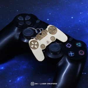 Può includere: Un portachiavi in legno a forma di controller PlayStation, con finitura in legno marrone e logo PlayStation nero. Il portachiavi è appoggiato su un controller PlayStation nero, su uno sfondo blu stellato.