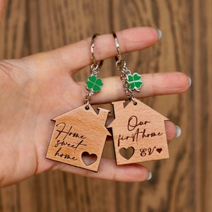 K&ouml;nnte beinhalten: Zwei Schl&uuml;sselanh&auml;nger aus Holz in Hausform mit dem Text "Home sweet home" und "Our first home &" eingraviert. Jeder Schl&uuml;sselanh&auml;nger hat einen kleinen gr&uuml;nen Gl&uuml;ckskleeblatt-Anh&auml;nger.