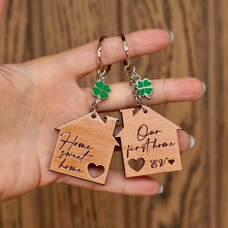 K&ouml;nnte beinhalten: Zwei Schl&uuml;sselanh&auml;nger aus Holz in Hausform mit dem Text "Home sweet home" und "Our first home &" eingraviert. Jeder Schl&uuml;sselanh&auml;nger hat einen kleinen gr&uuml;nen Gl&uuml;ckskleeblatt-Anh&auml;nger.