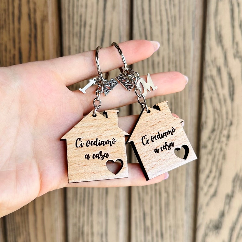 K&ouml;nnte beinhalten: Zwei Schl&uuml;sselanh&auml;nger aus Holz in Hausform mit dem Text "Ci vediamo a casa" (Wir sehen uns zu Hause) und einem Herz-Ausschnitt. Die Schl&uuml;sselanh&auml;nger sind an silbernen Schl&uuml;sselringen mit kleinen silbernen Anh&auml;ngern befestigt.