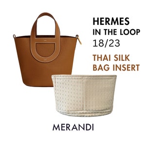 Pode incluir: Uma bolsa de couro marrom com uma alça curva e uma inserção de bolsa de seda tailandesa combinando. A bolsa tem um fecho dourado e o texto "HERMES IN THE LOOP 18/23 THAI SILK BAG INSERT" é visível.
