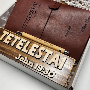 May include: A gift set featuring a brown leather journal with a strap closure, a wooden sign with the text "TETELESTAI John 19:30", and a bamboo pen. The journal and pen have the same text. The items are in a white box.