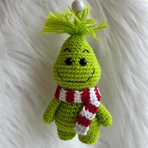 May include: A handmade green crochet ornament shaped like a character with a smiling face, black eyes, and a red and white striped scarf. The ornament has a green fringe on top and a white bead and green string for hanging.