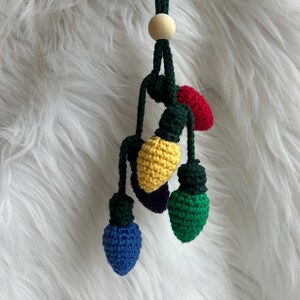 May include: A handmade crochet Christmas light ornament. The ornament features six colorful lights in red, yellow, green, blue, and purple, with green stems and caps. A wooden bead and dark green cord are at the top.