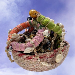 Op de afbeelding: Een geweven mand gevuld met kleurrijke kralen, schelpen en stofresten. De mand is gemaakt van natuurlijke vezels en heeft een rustieke, handgemaakte uitstraling.