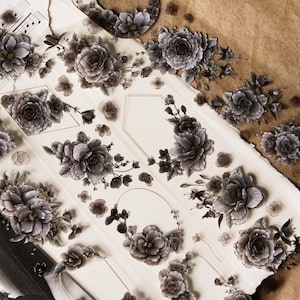 May include: Floral stickers with dark grey and black peony and rose designs. The stickers are arranged on a white background with torn edges and a brown surface. The floral elements include various sizes of flowers and leaves.