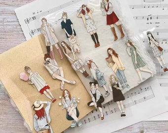 May include: A collection of stickers featuring illustrations of women in various outfits and poses. The stickers are in a clear plastic sleeve and are placed on top of sheet music and a brown paper envelope.