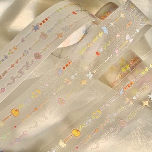 May include: Several rolls of decorative washi tape with a transparent base and colorful, glittery designs. The tape features repeating patterns of stars, bows, hearts, and other whimsical shapes in pastel colors. The tape is ideal for scrapbooking and crafts.
