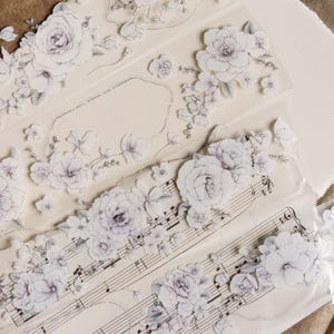 May include: Close-up of decorative floral washi tape rolls. The tape features white and grey flowers, including roses and smaller blossoms, with musical notes on some rolls. The background is a light beige.