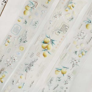 May include: Washi tape rolls with illustrations of lemons, white flowers, and greenery. The tape has a transparent background with text and small embellishments. The colour palette includes yellow, white, and light green.