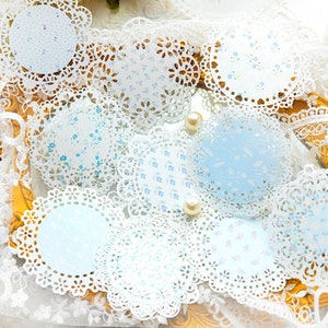 May include: A collection of round paper doilies with intricate lace edges. The doilies feature delicate floral patterns in shades of blue and white. Some doilies have a solid light blue background. Pearls are scattered around the doilies.