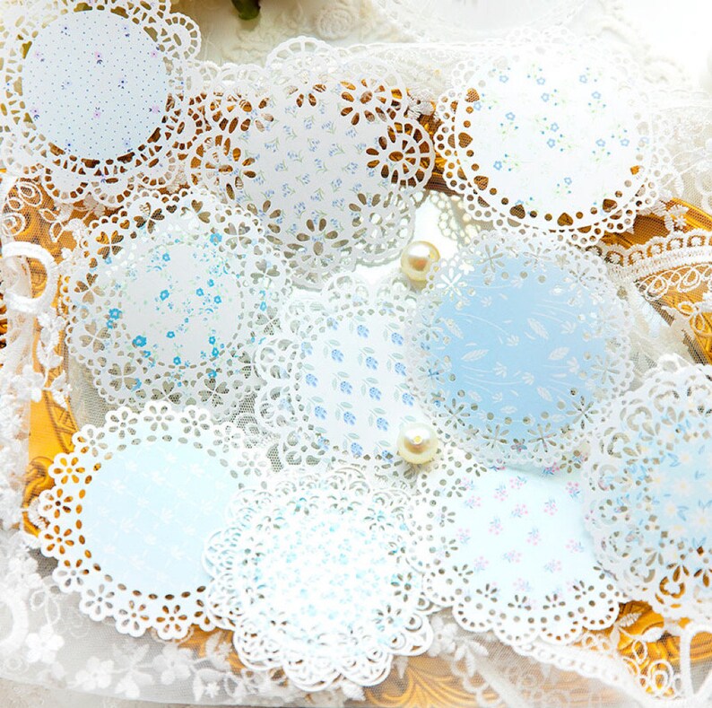 May include: A collection of round paper doilies with intricate lace edges. The doilies feature delicate floral patterns in shades of blue and white. Some doilies have a solid light blue background. Pearls are scattered around the doilies.