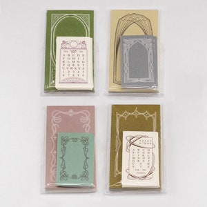 May include: Four sets of decorative stationery in clear plastic packaging. Each set includes a larger card with an ornate frame design and a smaller card with text and numbers. Colours include green, tan, pink, and olive.