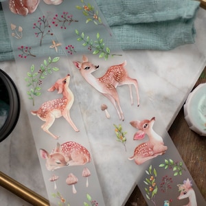 May include: Washi tape with watercolour illustrations of deer, mushrooms, and foliage. The tape has a translucent background and features various deer poses, including standing, lying down, and looking up. The colour palette includes soft browns, pinks, and greens.