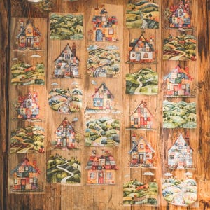 May include: A collection of colorful, illustrated house stickers on a wooden surface. Each sticker depicts a unique cottage with varying architectural styles and landscape scenes, featuring rolling hills and winding roads. The stickers are arranged in a grid pattern.