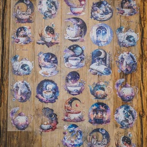 May include: A sheet of decorative stickers with celestial and tea party designs. The stickers depict teacups, teapots, moons, and whimsical illustrations in purple, blue, and white. The stickers are displayed on a wooden surface.