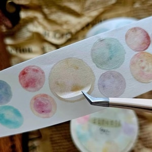 Könnte beinhalten: Ein Streifen dekorativen Washi-Tapes mit Kreisen im Aquarellstil in Pastellfarben. Eine Pinzette hebt einen Kreis vom Band ab. Das Band befindet sich auf einer Oberfläche mit unscharfem Hintergrund.