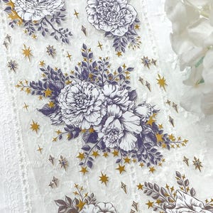 May include: Decorative floral tape featuring white peony flowers with purple accents and gold star embellishments. The tape has a delicate, transparent appearance, ideal for scrapbooking and craft projects.