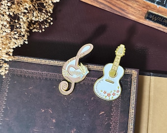 May include: Two musical-themed pins. One is a treble clef with a bird and musical notes, the other is a light blue guitar with floral accents. Both pins have gold-coloured outlines and are on a dark brown surface.