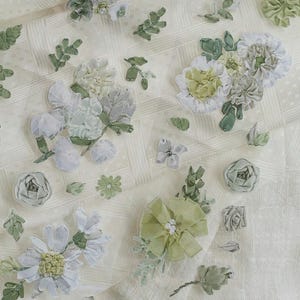 May include: Close-up of cream fabric decorated with handmade floral embellishments. The design includes flowers in white, green, and grey, with detailed textures. The overall aesthetic is soft and elegant, with a focus on the craftsmanship.