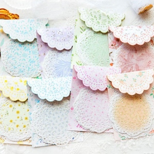May include: A collection of colorful, patterned paper envelopes with scalloped edges and matching doilies. The envelopes feature floral and lace designs in pastel colors, including pink, yellow, and blue. A gold-framed picture and a magnifying glass are also visible.
