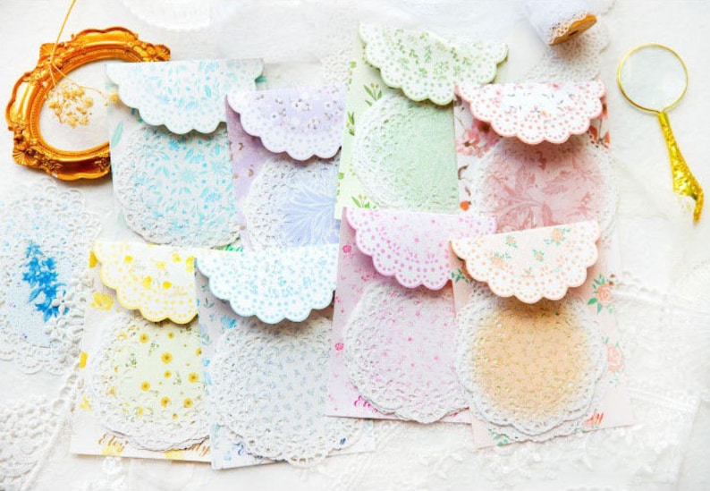 May include: A collection of colorful, patterned paper envelopes with scalloped edges and matching doilies. The envelopes feature floral and lace designs in pastel colors, including pink, yellow, and blue. A gold-framed picture and a magnifying glass are also visible.