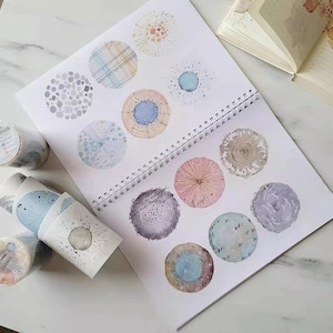 May include: A spiral-bound sketchbook open to a page of watercolor circle designs in various colors and patterns. Rolls of matching washi tape are visible on the left. The designs include blue, pink, and beige circles with textured and patterned details.