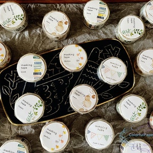 May include: A selection of masking tape rolls featuring diverse patterns, such as floral, geometric, and book designs. The tape rolls are displayed on a dark tray with a gold edge. The labels display "masking tape" and Japanese text.