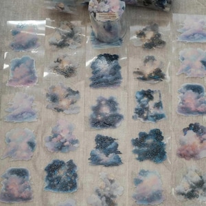 May include: A roll of clear adhesive tape featuring a repeating pattern of individual cloud stickers in shades of blue, purple, pink, and white. The stickers depict various cloud formations with a celestial, starry texture throughout the designs.