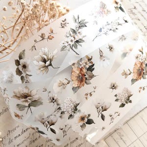 May include: Decorative floral washi tape featuring a repeating pattern of watercolor flowers in shades of white, brown, and peach. The tape is translucent and appears to be rolled up, with the floral design visible on the surface.
