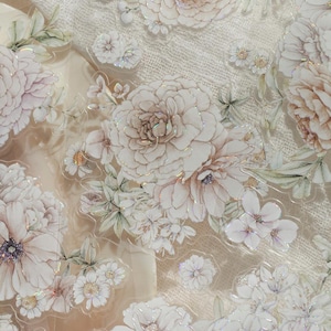 May include: Close-up of floral stickers with white, cream, and light brown flowers. The stickers have a glittery, iridescent finish and are arranged on a textured, off-white background. The flowers include peonies, daisies, and other blossoms.