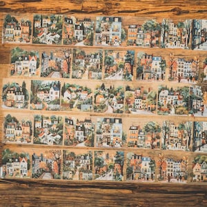 May include: Decorative washi tape featuring a repeating pattern of quaint European townhouses and streets. The illustrations include trees, people, and various architectural styles, with a warm, inviting color palette. The tape is arranged on a wooden surface.