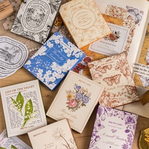 May include: A selection of decorative cards with floral and botanical designs. The cards feature various colours, including blue, beige, and purple, with text and illustrations of flowers, butterflies, and botanical elements. Some cards have text that reads "Lily of the Valley" and "Wedding".