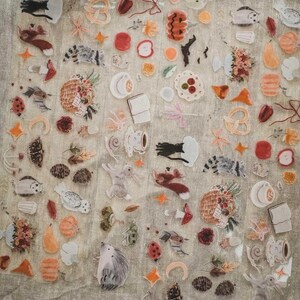 May include: A collection of clear stickers featuring various illustrations. The stickers include images of animals like rabbits, raccoons, and hedgehogs, along with food items such as apples, pretzels, and teacups. There are also floral and nature-themed designs.