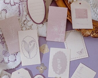 May include: A collection of stationery items, including tags and cards, in shades of lavender and cream. The designs feature floral illustrations and delicate line drawings. Some cards have lines for writing. The overall aesthetic is vintage and romantic.