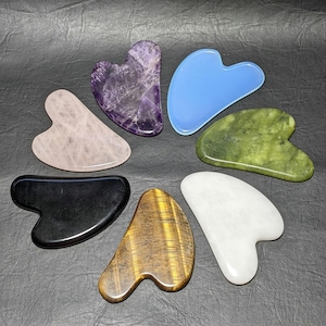 Peut inclure: Ensemble de sept outils de gua sha en forme de cœur, faits de différents types de pierres, notamment l'améthyste, le quartz rose, le jade, l'obsidienne noire, l'œil de tigre et le quartz blanc. Les outils sont disposés en cercle sur une surface gris foncé.