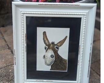 May include: A framed watercolor painting of a donkey with a curious expression. The donkey is brown and white with a black nose and eyes. The painting is set in a white frame with a black mat.